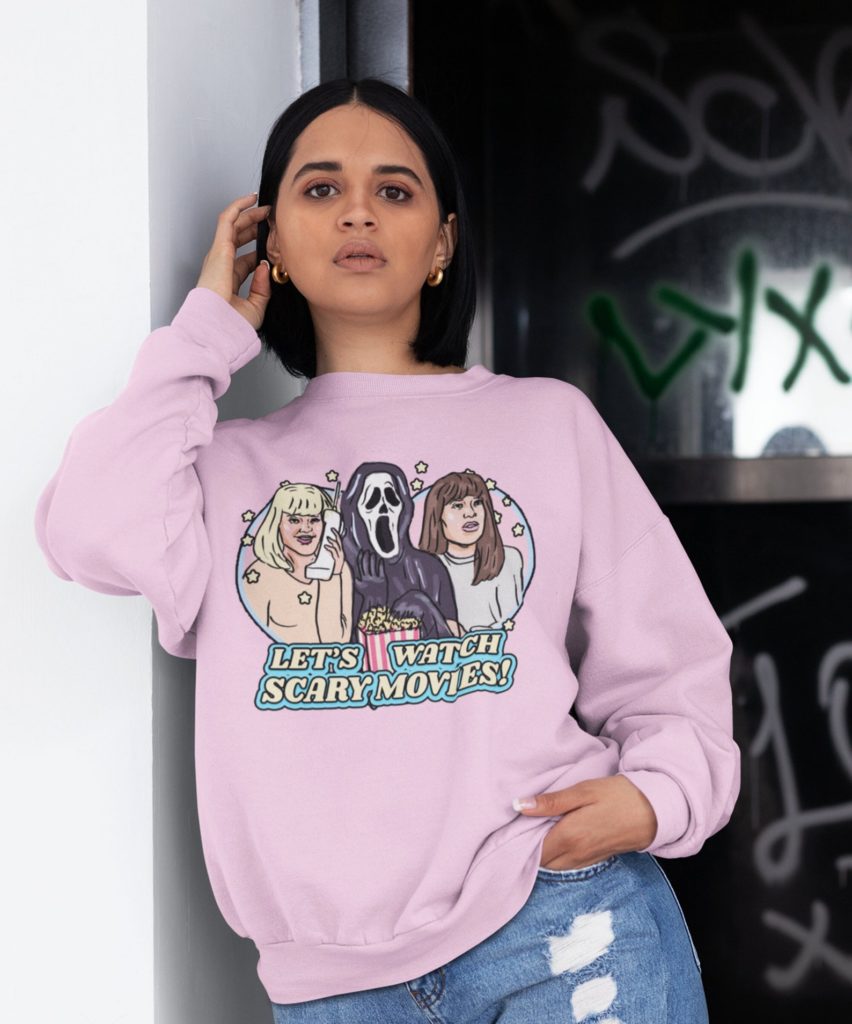 Photo of female model wearing pastel pink crew neck sweatshirt with retro illustration of Casey Becker (Drew Barrymore), Sidney Prescott (Neve Campbell), and Ghostface eating popcorn together with text saying "Let's Watch Scary Movies!" below