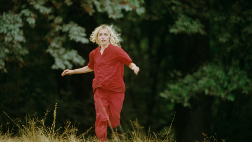 Movie still from Strange Darling (2024) of Willa Fitzgerald's character wearing red scrubs and running in fear through a field